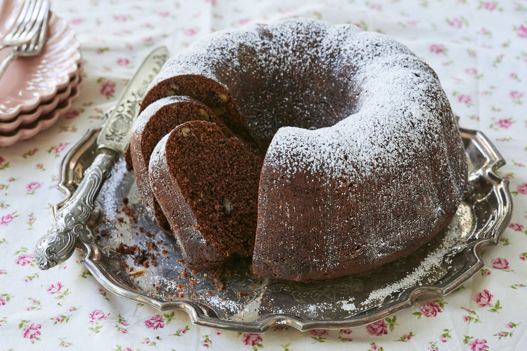 A homemade applesauce cake, baked in a bundt pan, is served on a silver tray. The cake has been sliced into, displaying bits of walnuts inside.