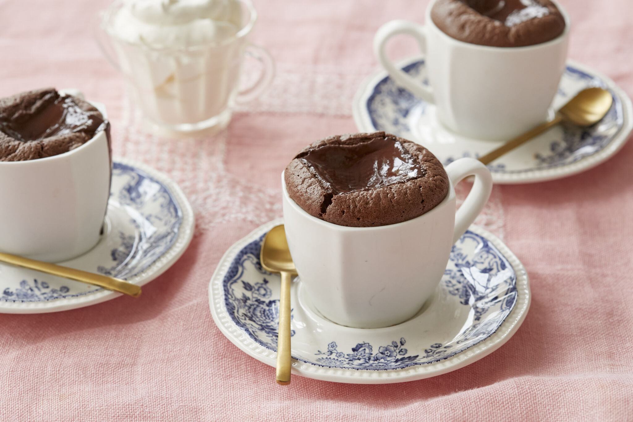 Baked hot chocolate is served in two white mugs on blue floral saucers with golden spoons. A glass cup of whipped cream is on the side. It's both light and rich, with an addictive contrast between the melty center and the surrounding soufflé-like cake.