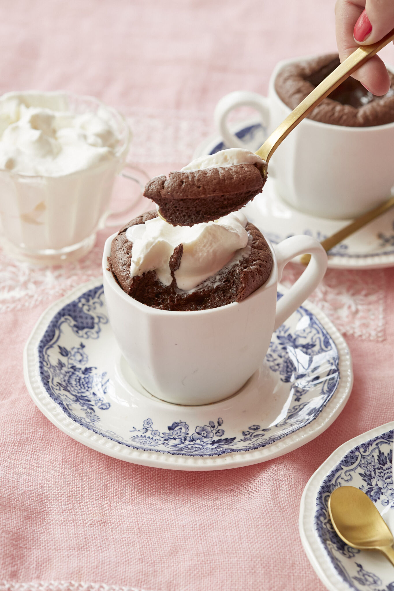 Baked hot chocolate is served in two white mugs on blue floral saucers with golden spoons. A glass cup of whipped cream is on the side. It's both light and rich, with an addictive contrast between the melty center and the surrounding soufflé-like cake. One mug of the baked hot chocolate is topped with softly whipped cream. One spoonful of scooped baked chocolate shows the moist interior. 