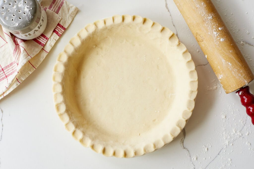 The Ballymaloe Creamed Butter Pie Crust is assembled into a pie pan with fluted edge. The flour shaker is in the top left corner, the wooden rolling pin with red handles is on the right side.
