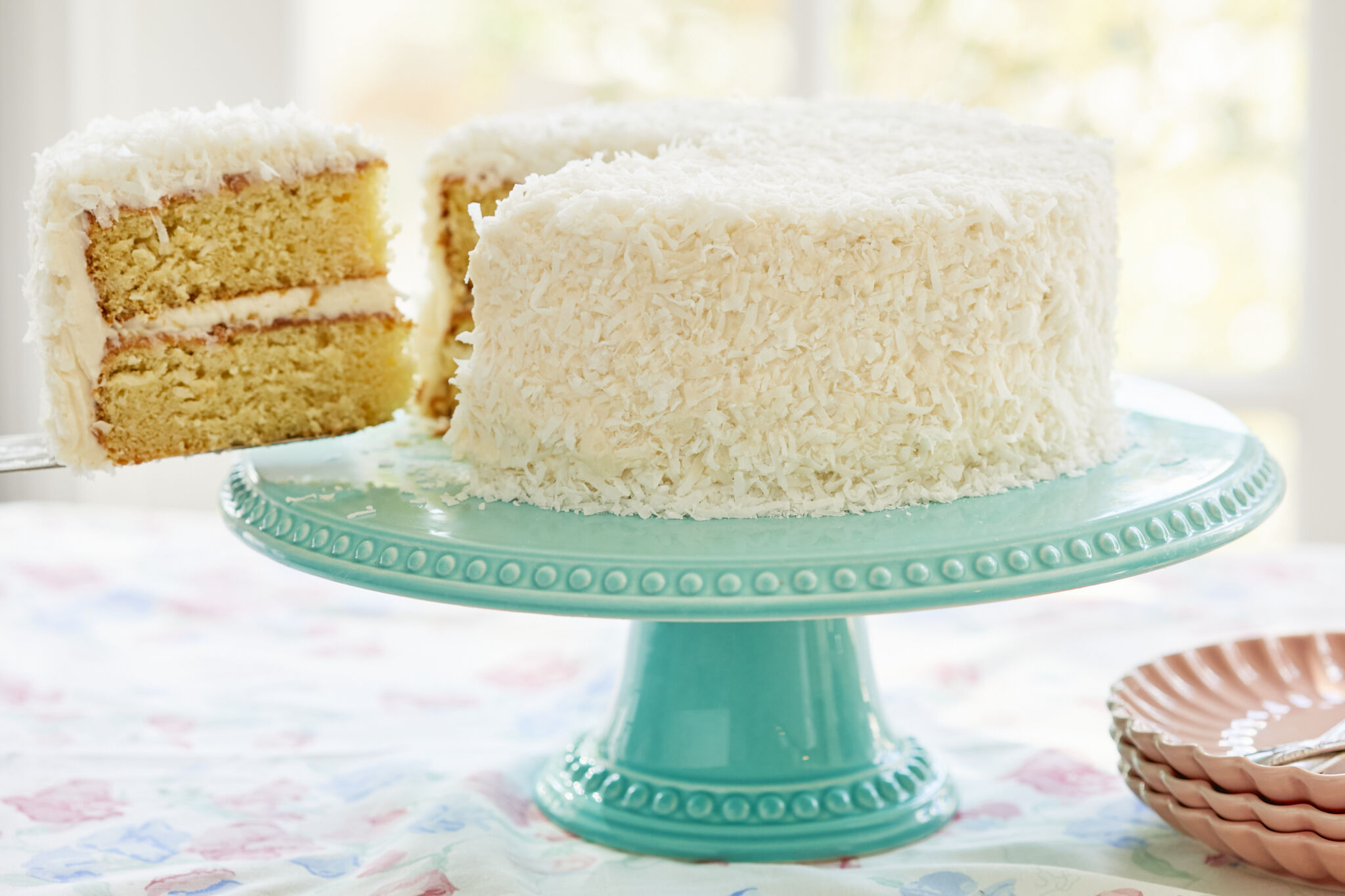 A slice was cut from and being lifted off the perfectly baked two-layer moist coconut cake, with white frosting between layers and on the outside, covered with crunchy coconut.