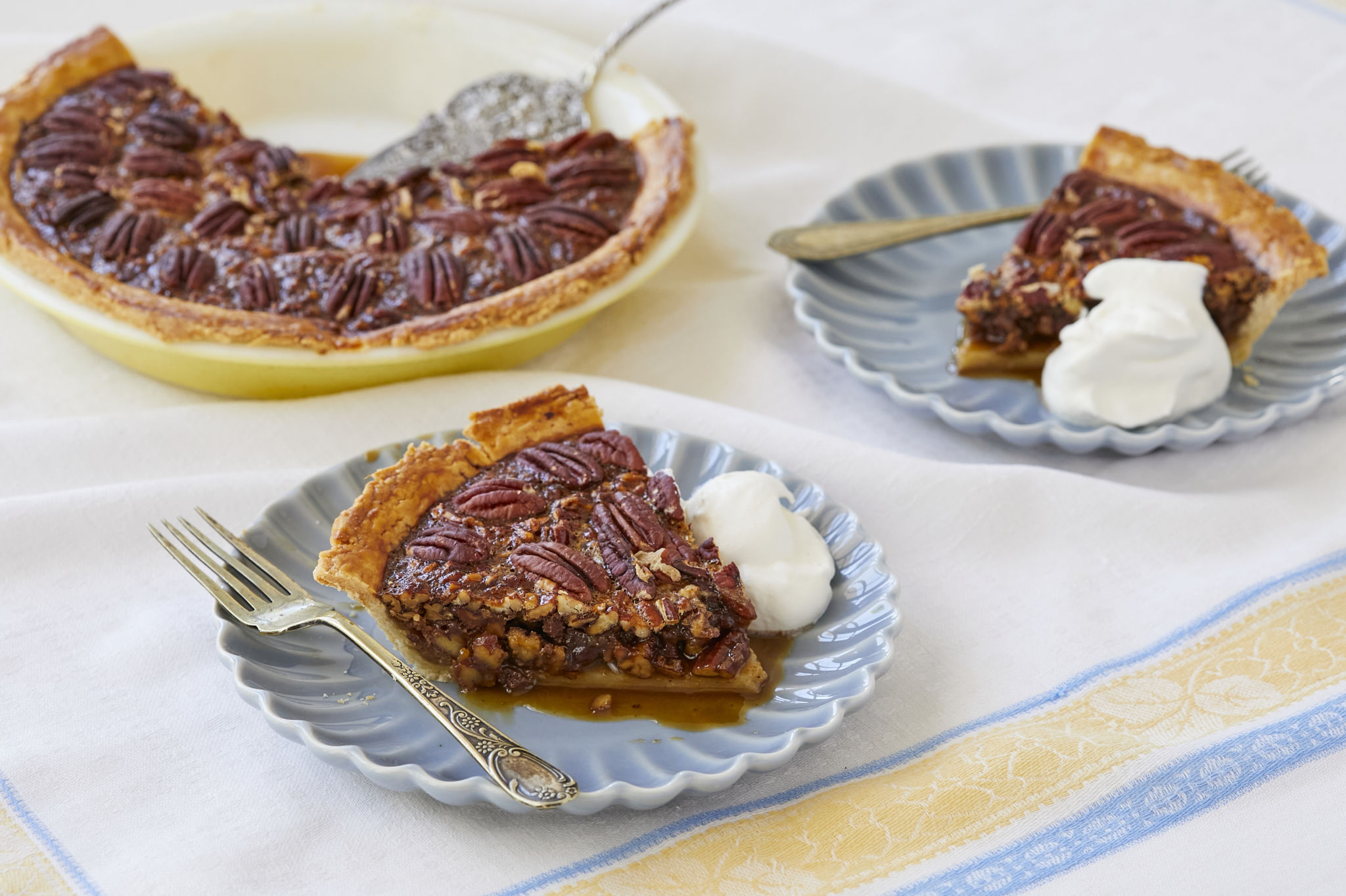 Two slices of homemade pecan pie are served on blue dishes next to the pie tin. There is fresh homemade whipped cream served with the pies.
