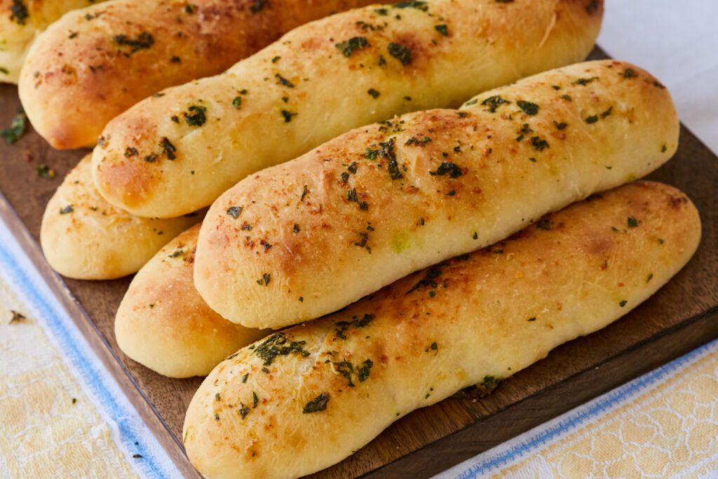 Garlic Breadsticks are shaped into long, thin rods and were baked until golden brown. The exterior looks slightly crispy with brushed butter, garlic and herbs.