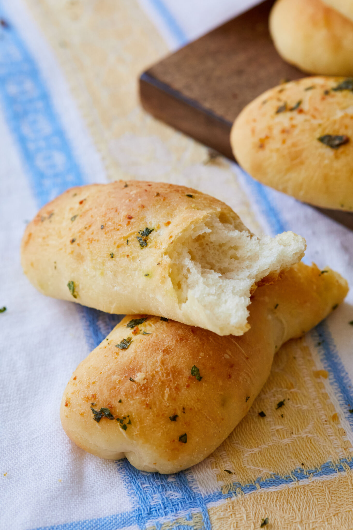Garlic Breadsticks are shaped into long, thin rods and were baked until golden brown. The exterior looks slightly crispy with brushed butter, garlic and herbs. One stick is broken into halves, showing the light fluffy crumb. 