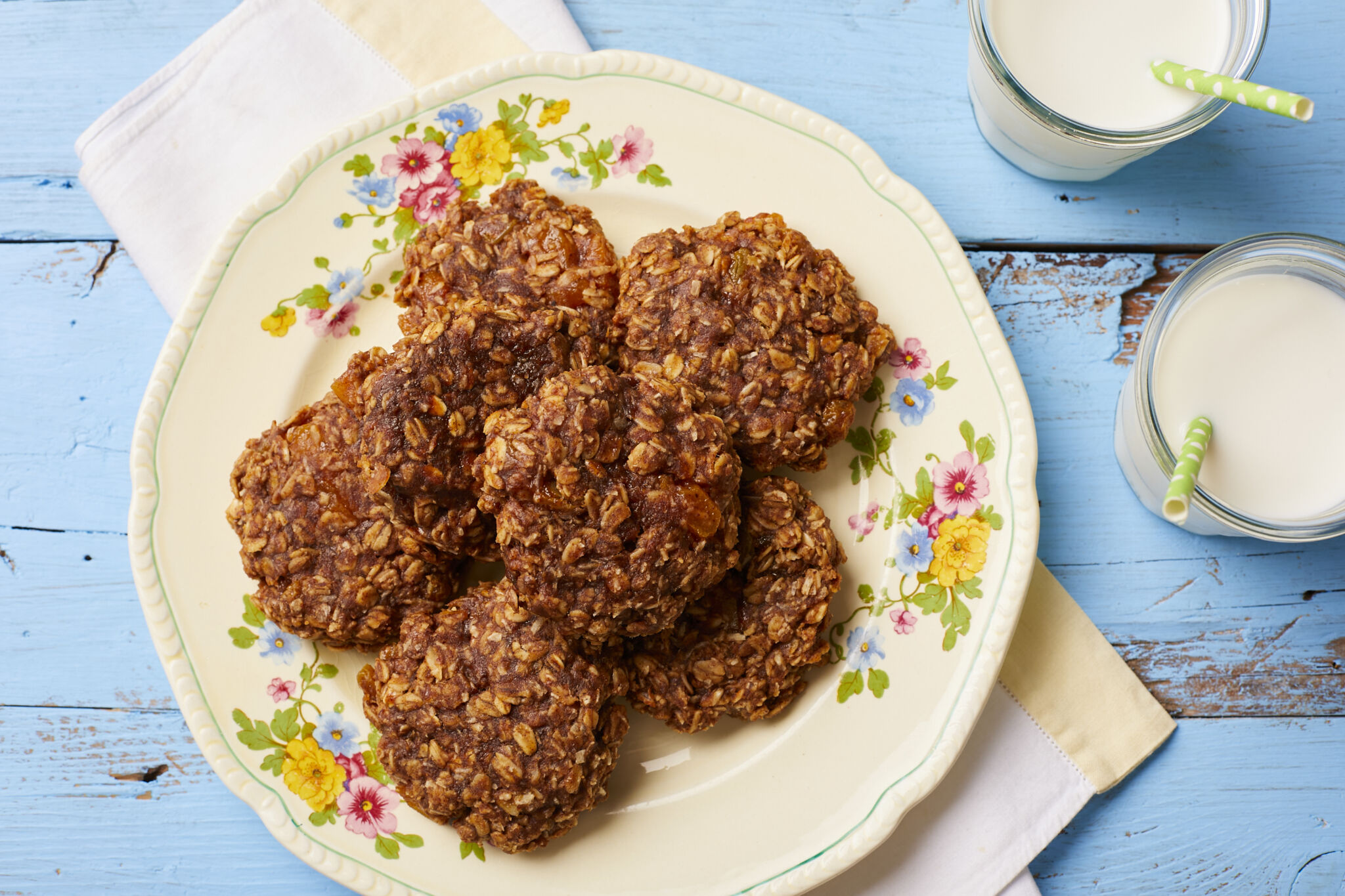 Golden brown breakfast cookies are served on a floral plate with two glasses of milk on the side with straws. Cookies are packed with delicious and nutritious rolled oats, dried fruit and coconut flakes.