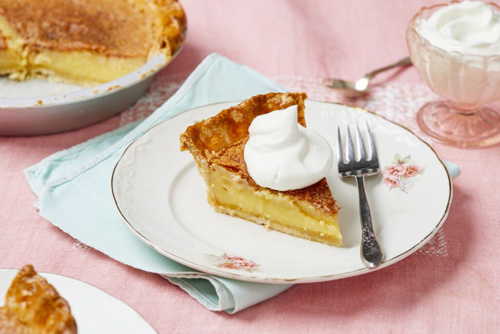 Two slices of the Classic Chess Pie are served on golden-rim dessert plates. The slice has flaky crust, silky filling, and crispy caramelized topping crowned with a big dollop of whipped cream. The rest of the pie is in the pie pan on the side. Extra whipped cream is served in a pink great depression glass cup.