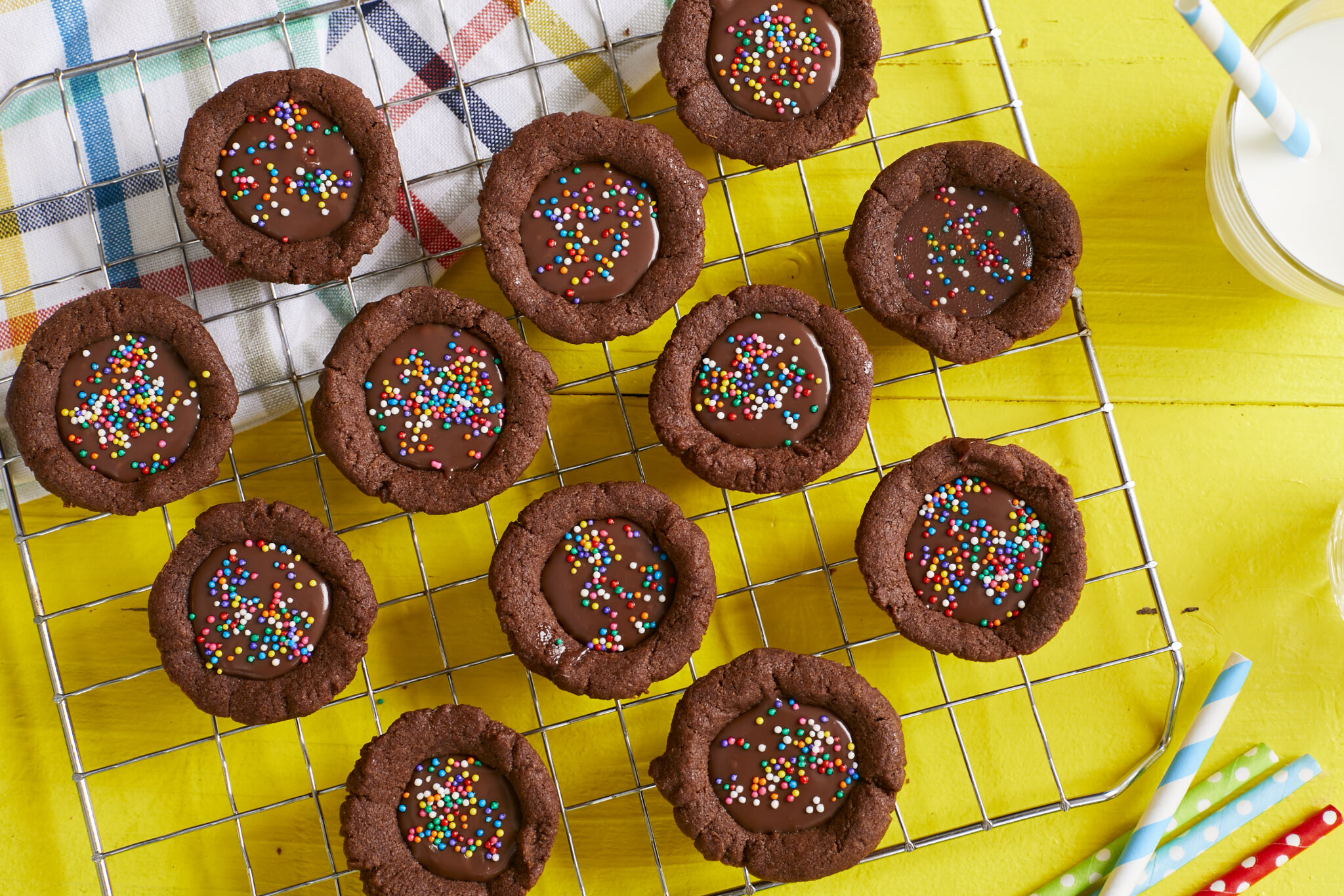 Bite-size Chocolate Cookie Cups are placed on a cooling rack. They are in signature chocolate brown color, have lightly crispy edges, soft and chewy inside with a silky ganache filling. They're decorated with colorful sprinkles. A glass cup of milk is served on the side.
