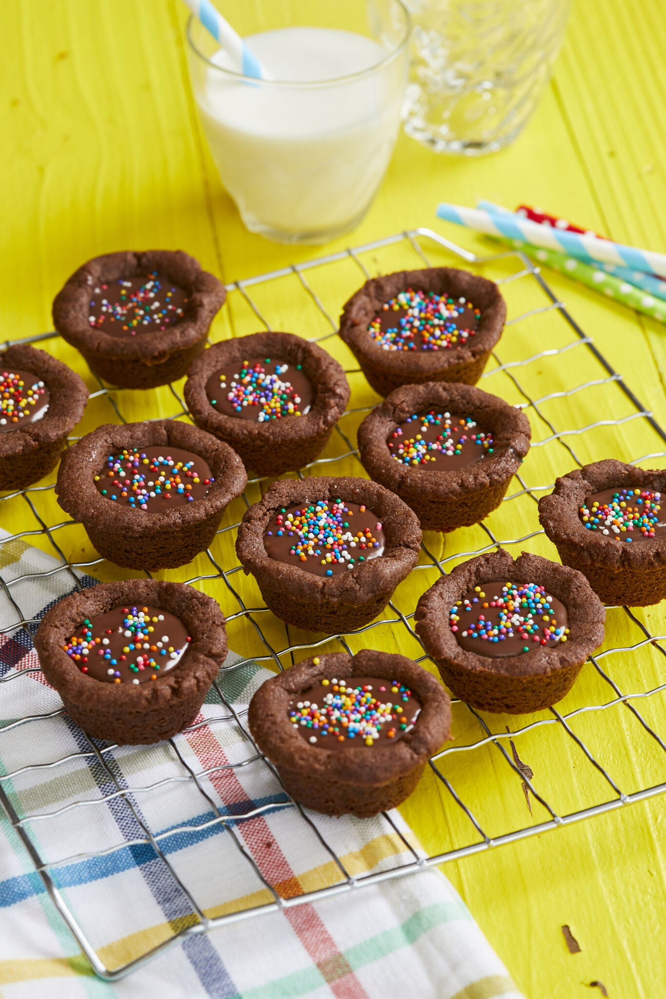 Bite-size Chocolate Cookie Cups are placed on a cooling rack. They are in signature chocolate brown color, have lightly crispy edges, soft and chewy inside with a silky ganache filling. They're decorated with colorful sprinkles. A glass cup of milk is served on the side. 
