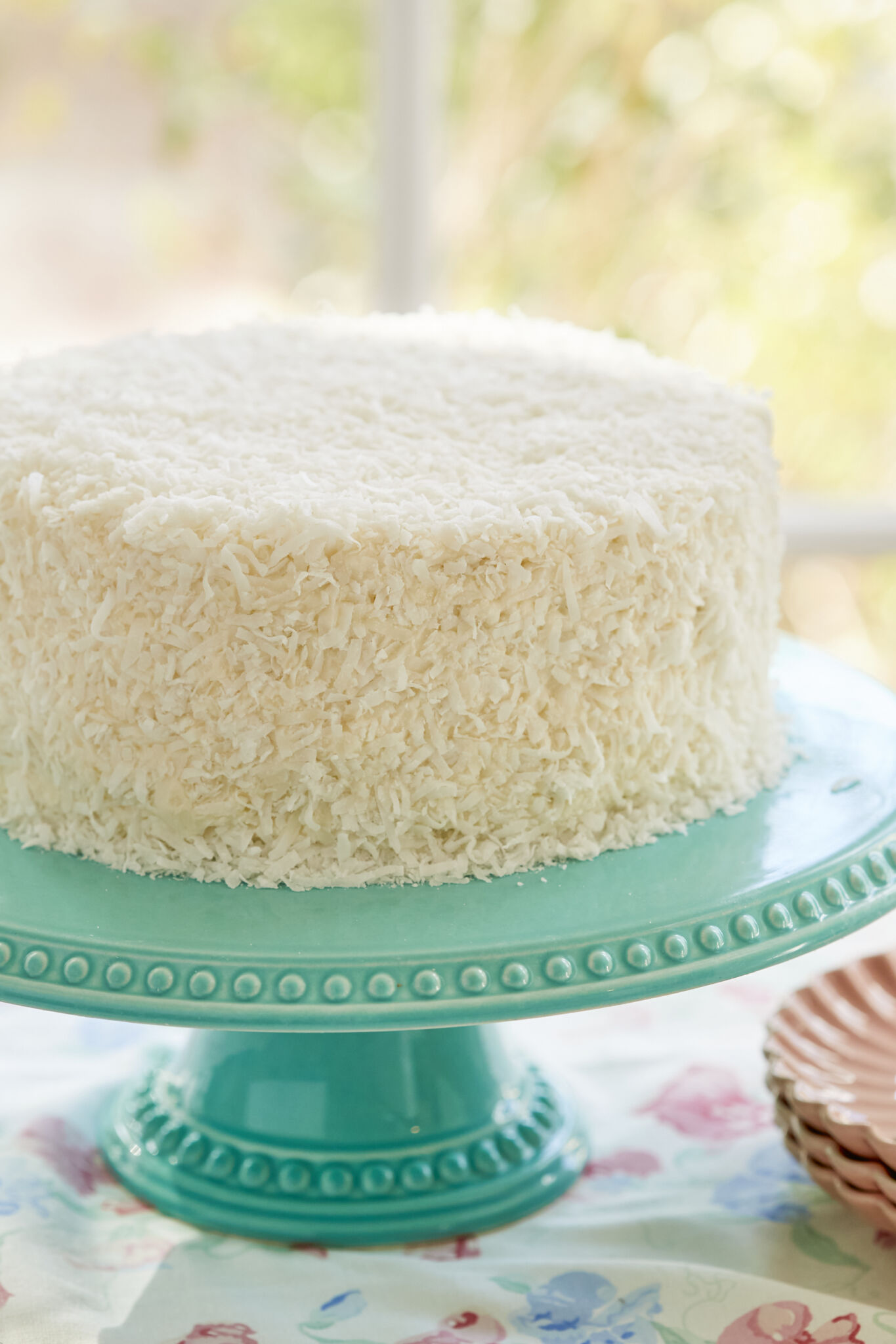 Incredibly Moist Coconut Cake covered with smooth frosting and crunchy coconut, on an aqua color cake stand. 