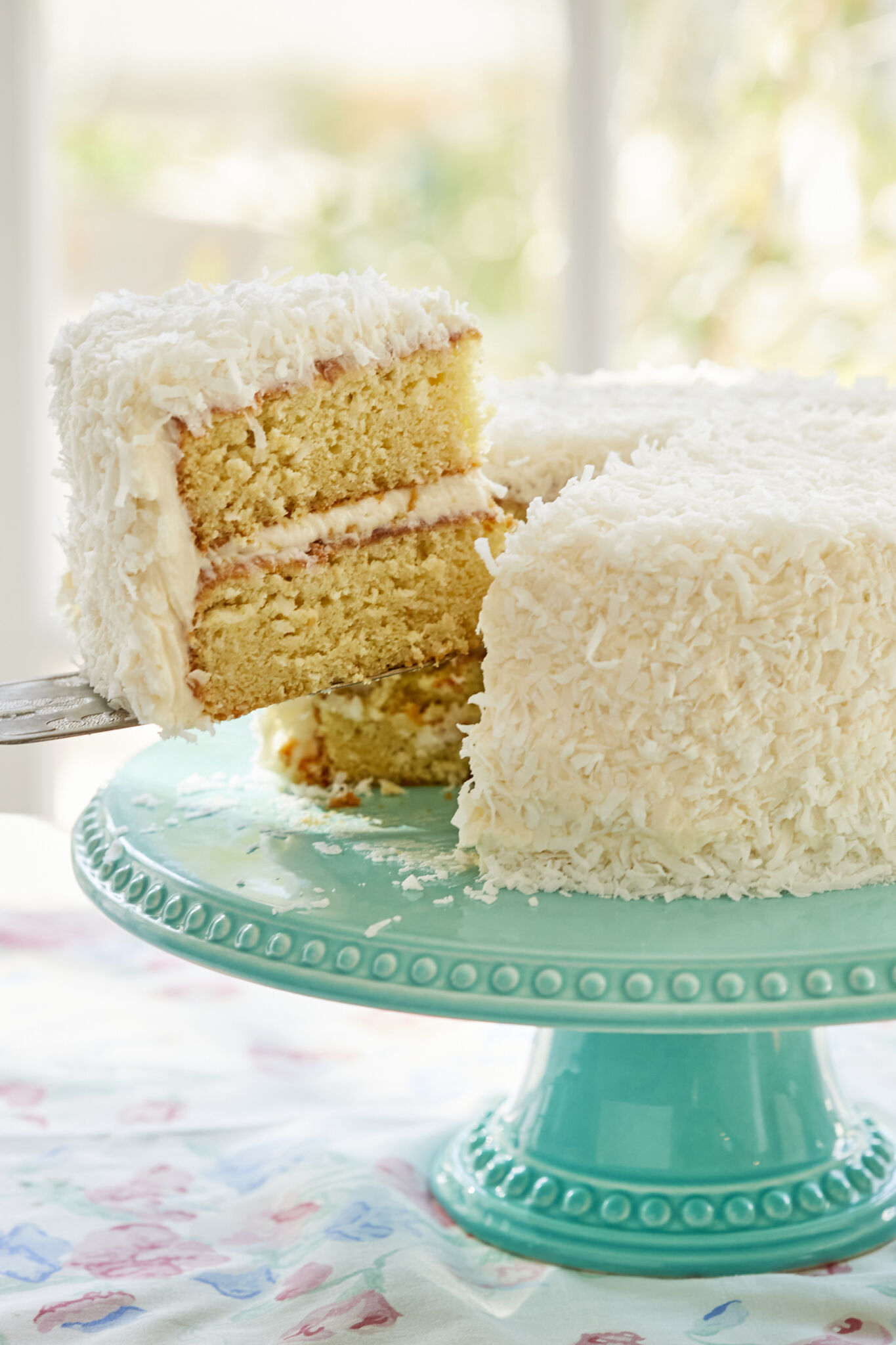 A slice was cut from and being lifted off the perfectly baked two-layer moist coconut cake, with white frosting between layers and on the outside. Topped with crunchy coconut.