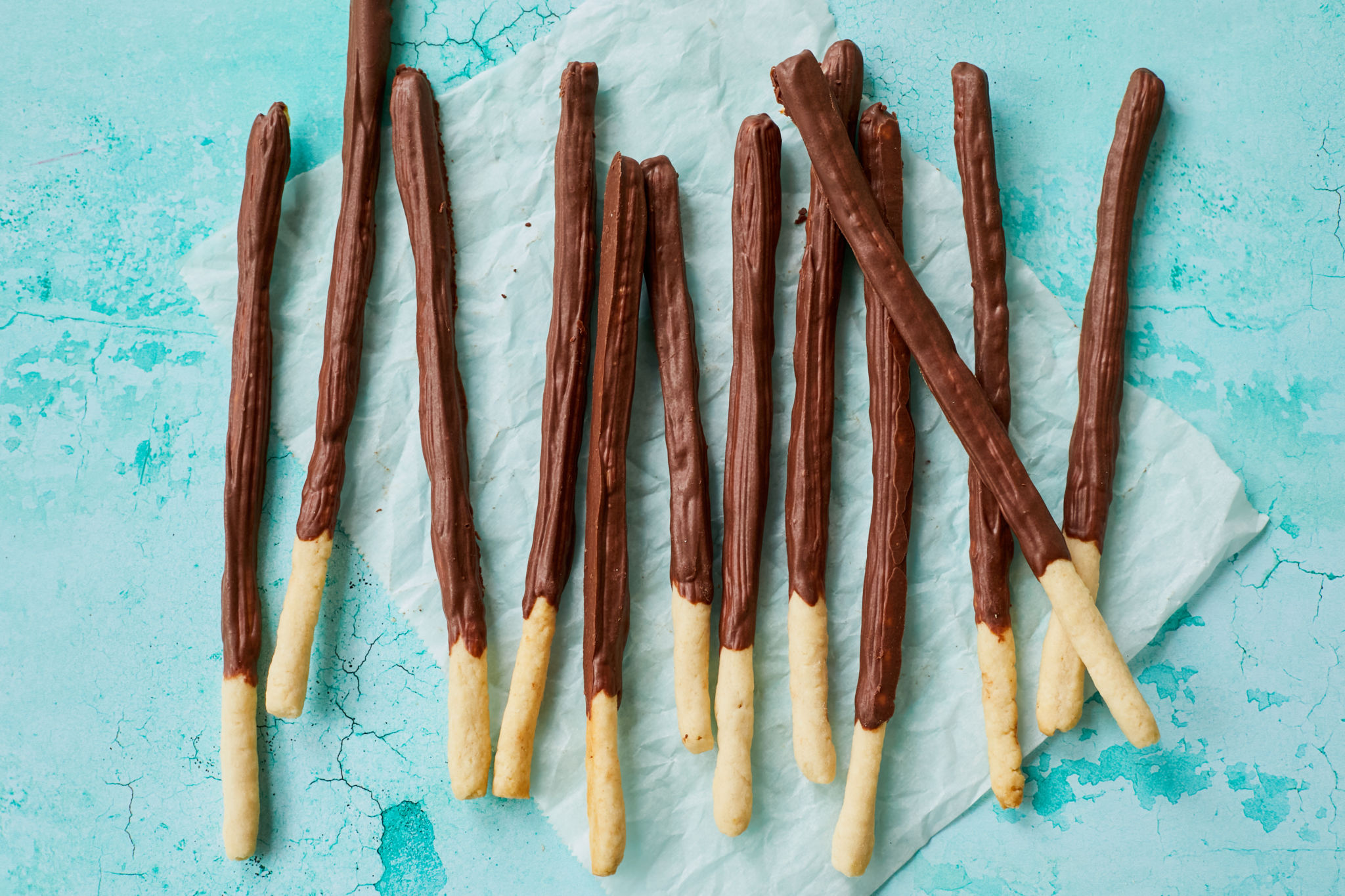 Homemade Pocky sticks are in a thin elongated-stick shape, with most of the stick covered in a chocolate-flavored coating while leaving a small section uncoated for easy handling. The cookies are placed in a glass Ball mason jar with a light teal background. On Pocky biscuit is being taken out of the jar.