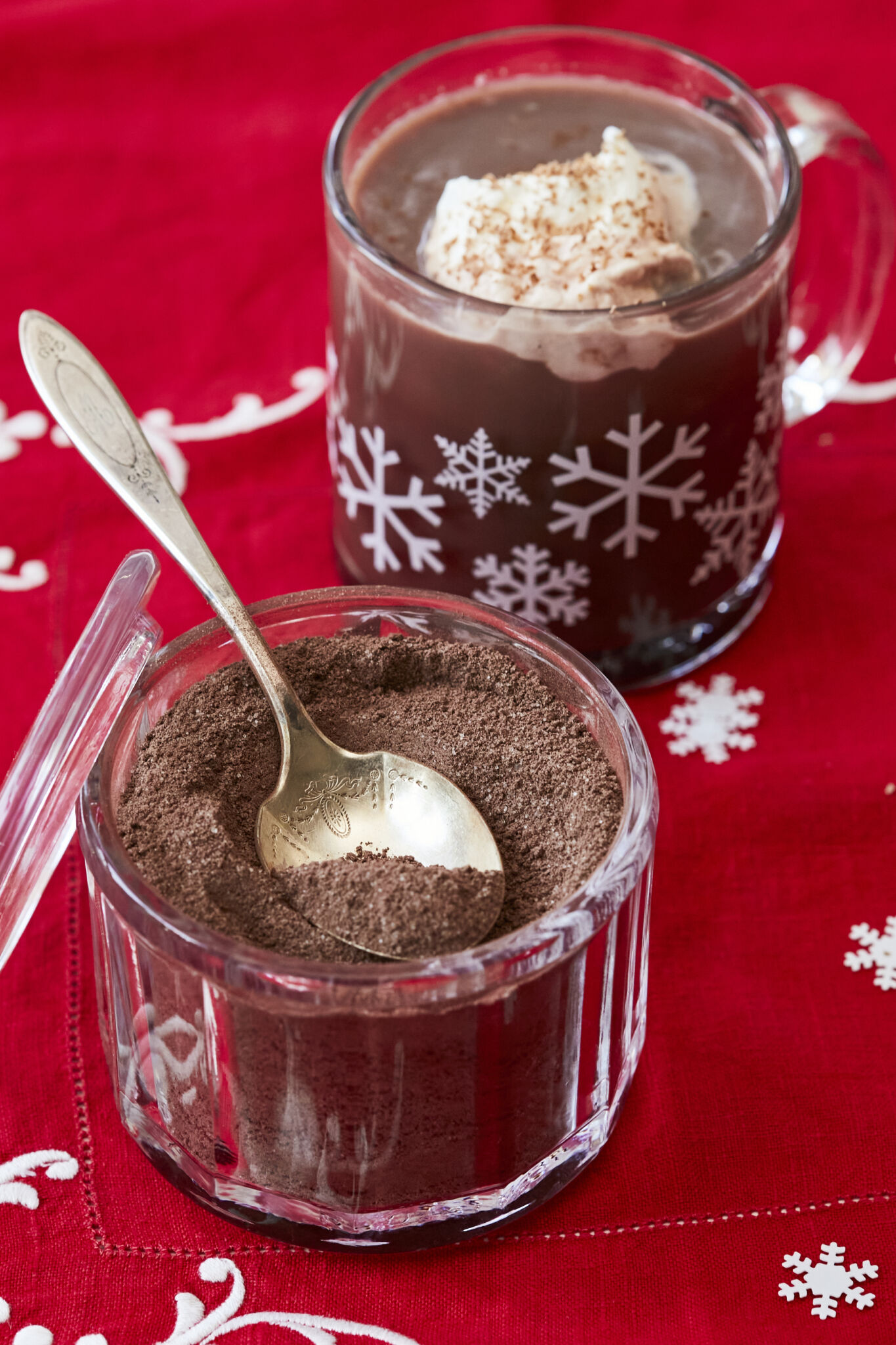 Homemade Hot Chocolate Mix in a glass jar with a golden spoon. A mug of hot chocolate made from this is next to it with whipped cream on top. 