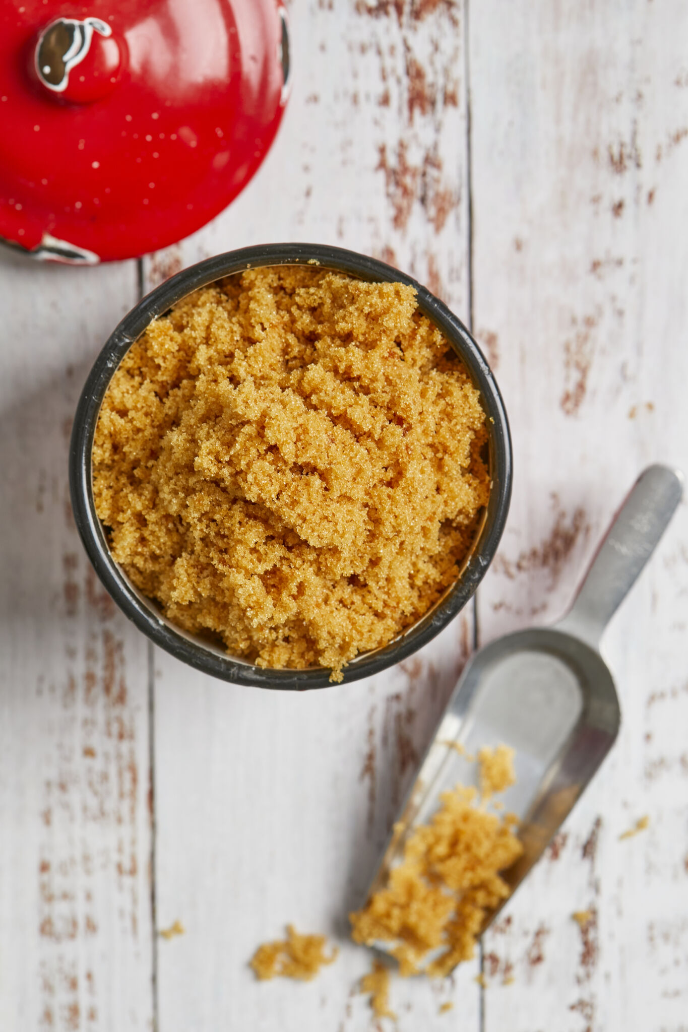 Homemade brown sugar is stored in an enamel mug with a lid in the top left corner and a metal scoop next to it in the bottom right corner.