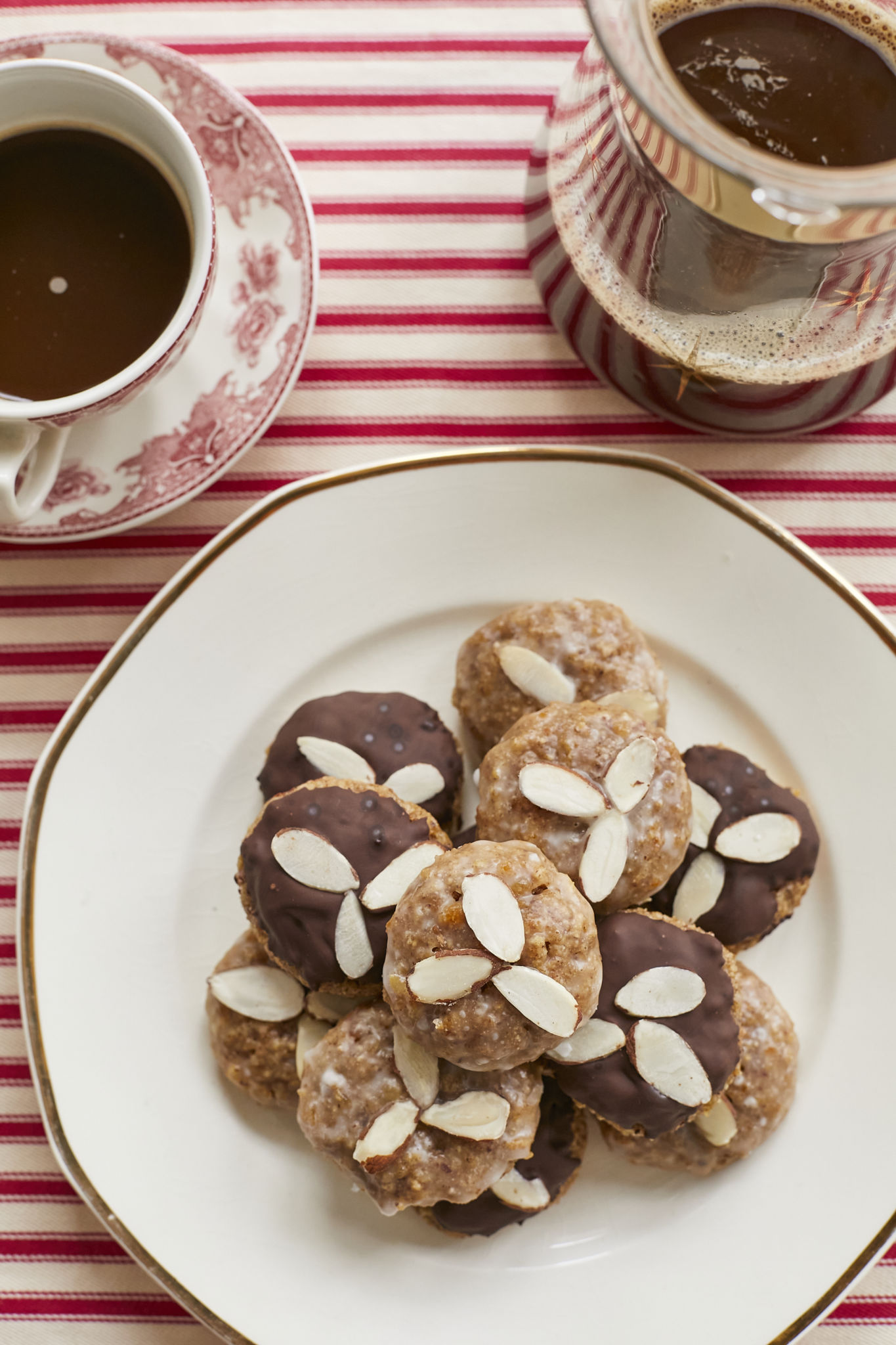 On a red and white table cloth, a white plate with half chocolate, half glazed Lebkuchen, covered in almonds, are served on a white plate next to two tea cups filled with thick hot chocolate.