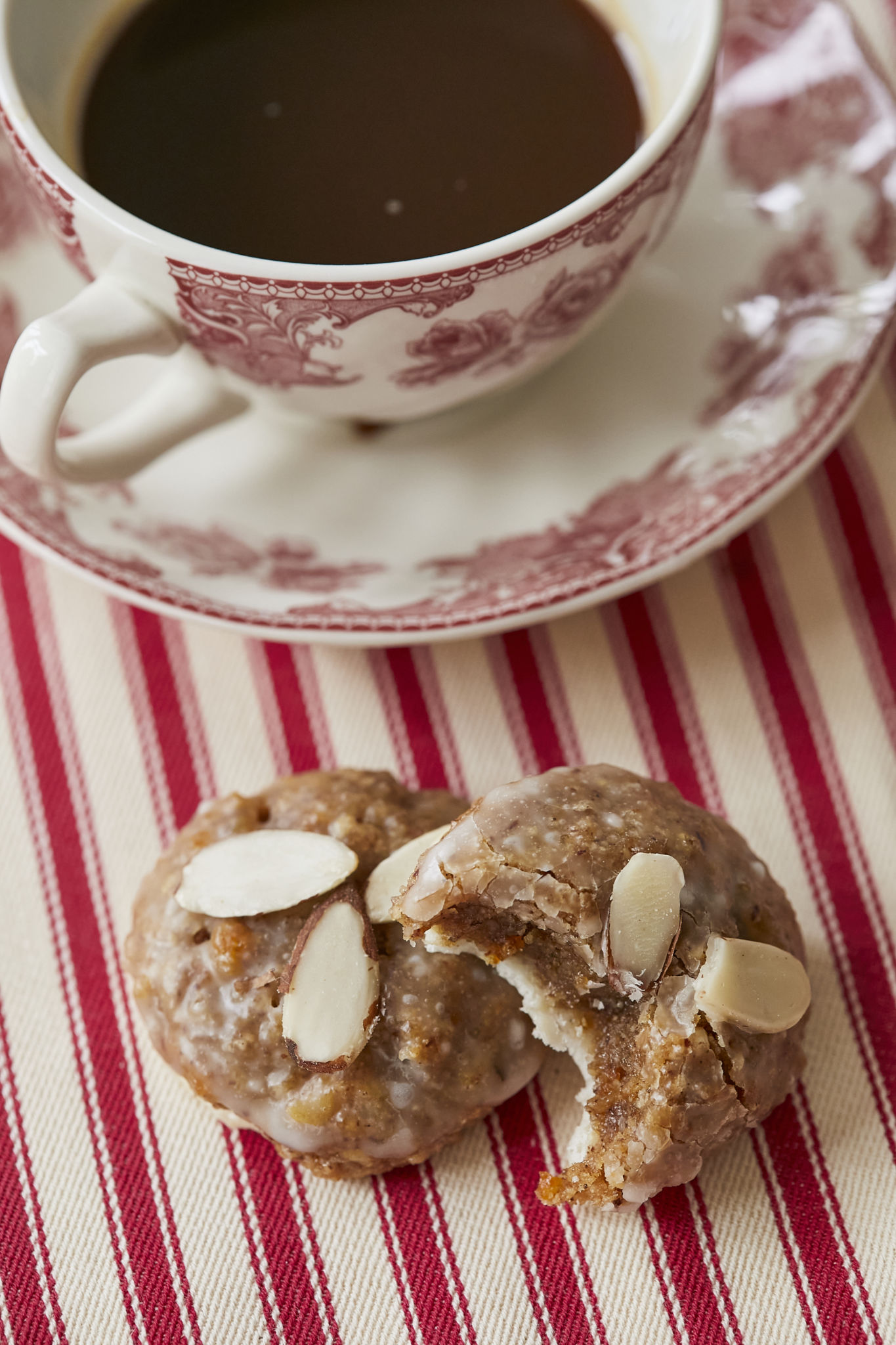 A Lebkuchen with a bite taken out of it show the different layers and textures of the cookie. On the bottom of the Lebkuchen is a edible paper wafter, the cookie itself looks chewy and packed with nuts, and the top is covered in a homemade glaze and slivered almonds. The two cookies are next to a tea cup with coffee on a red and white striped table cloth. 