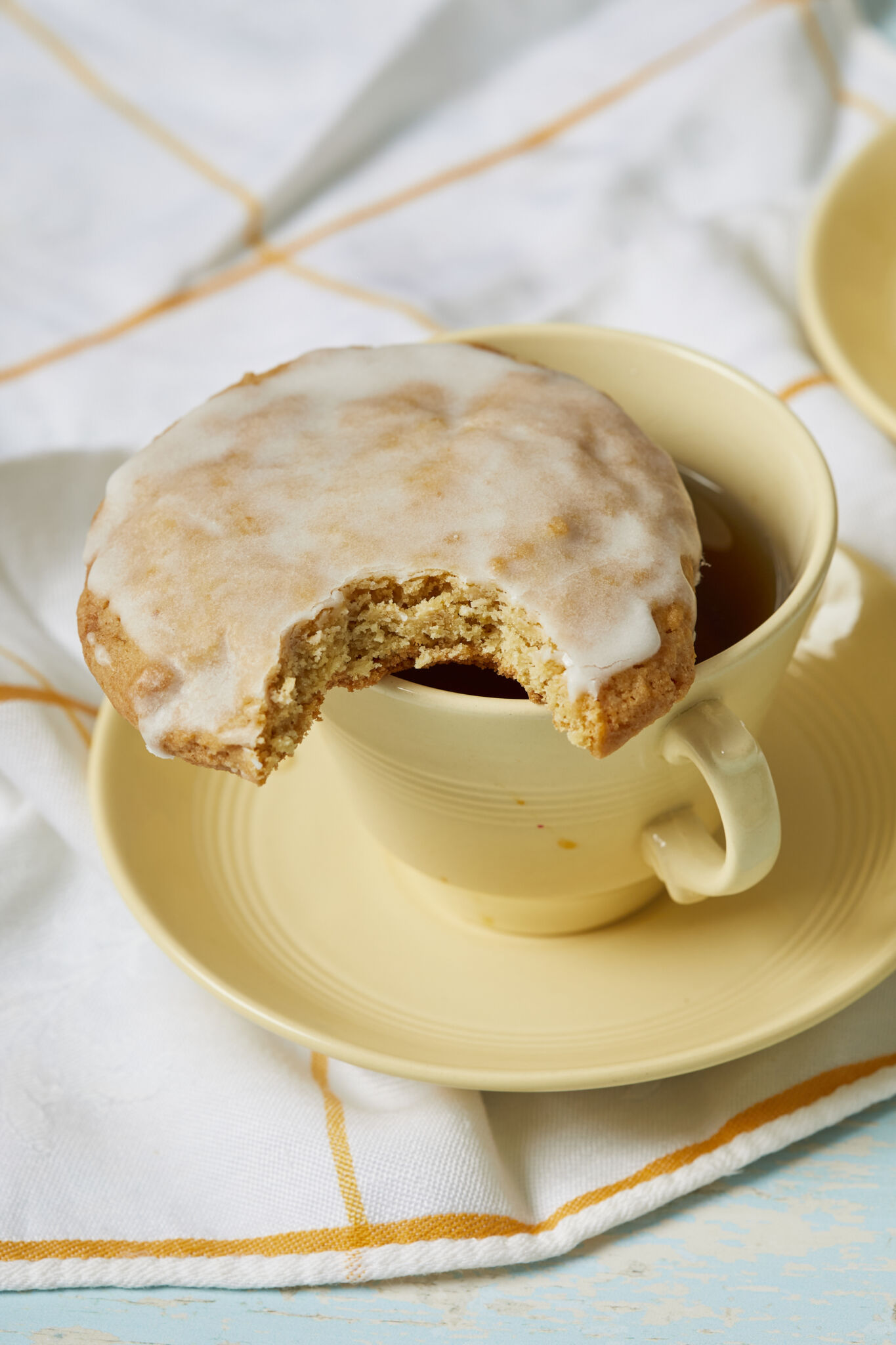 A soft lemon cookie covered with zesty lemon glaze, sitting on the rim of a tea cup, with one bite taken. 