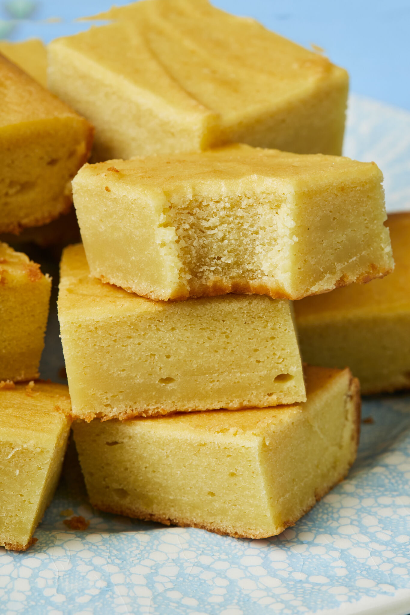 A close shot at a pile of golden luscious Mango Butter Mochi on a light teal-blue plate. A bite has taken from the front piece, showing the silky soft texture on top and crispy crust at the bottom.