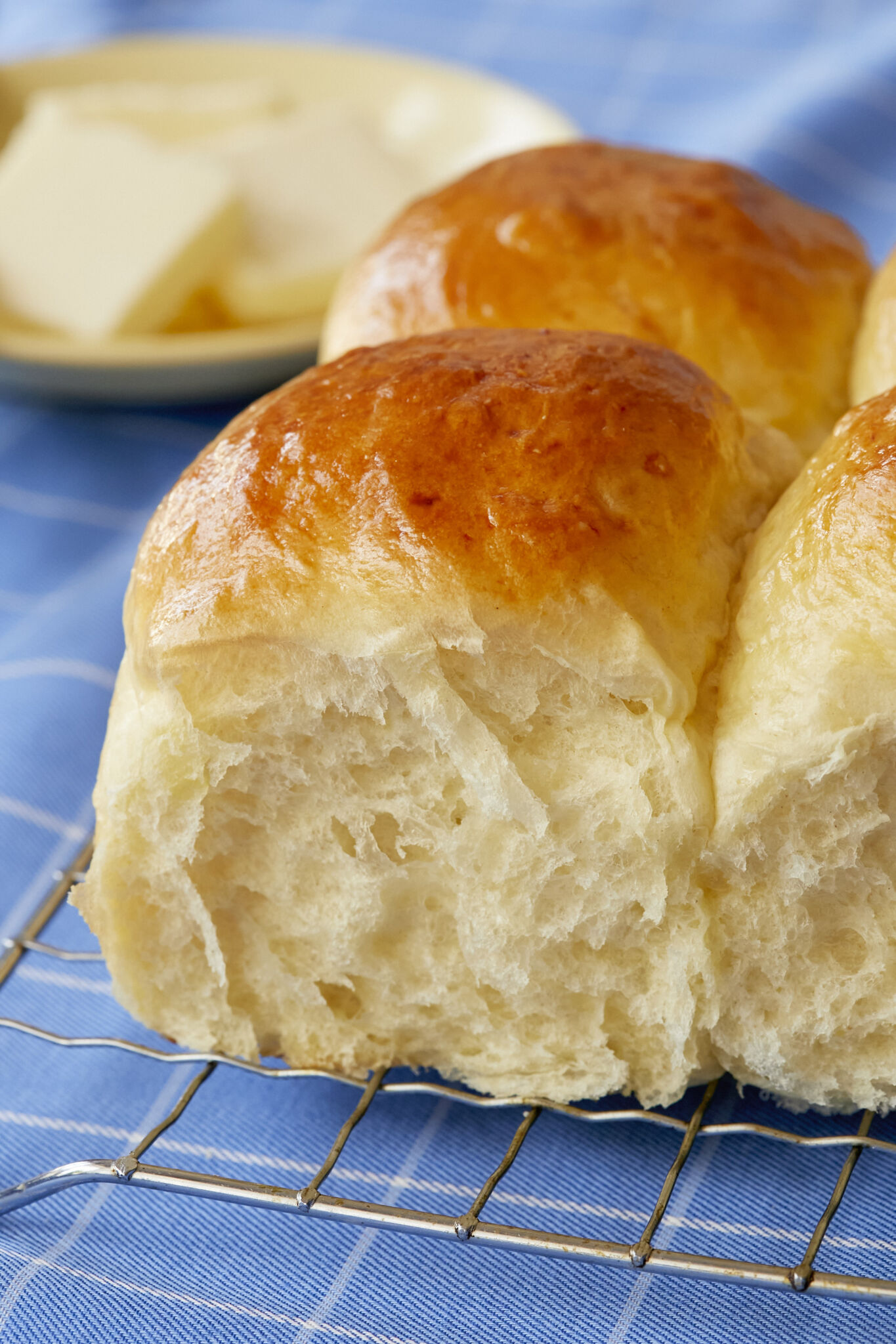 A close shot at Mashed Potato Dinner rolls which are cooling or a wire rack shows their soft and bubbly crumb and golden brown top. The rolls are served with butter on a small plate. 