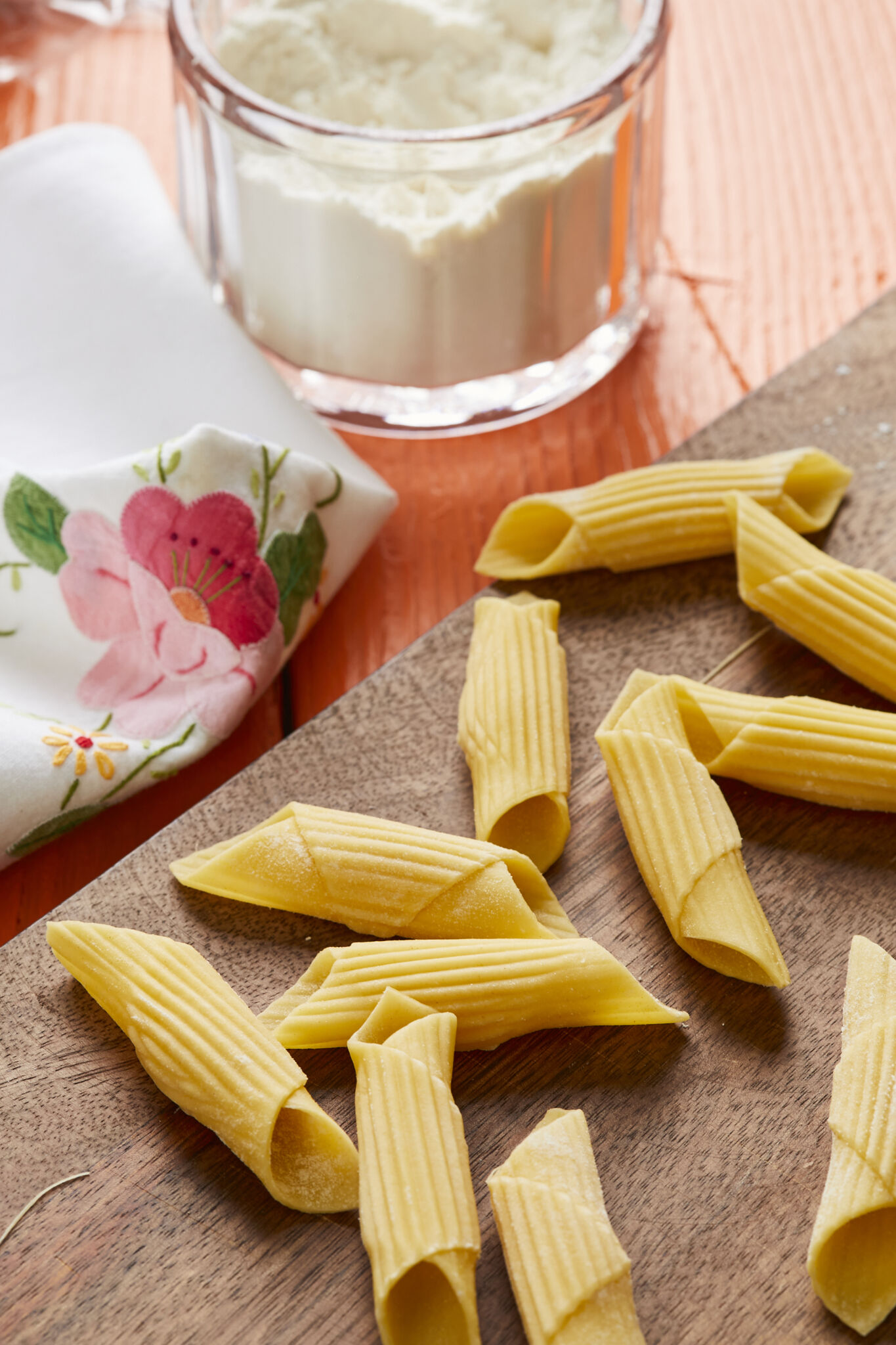 Penne pasta is drying on a wooden board. It's an eggless tubular-shaped pasta, cut diagonally on the ends and resembles an old-fashioned writing quill that was dipped in ink. A small glass bowl of flour is on the side.