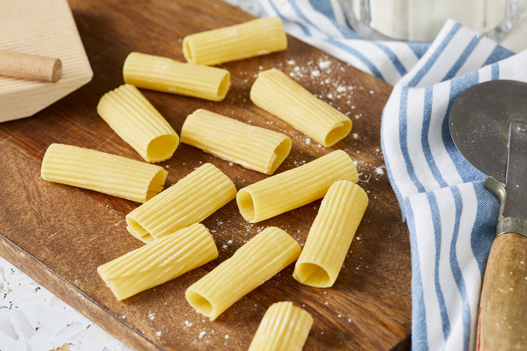 Rigatoni pasta is drying on a wooden board. It's in ridged, tubular shape and about 2 inches long with squared-off ends. It got the yellow color from semolina flour. A gnocchi board is on the left and a pasta cutter is on the right.