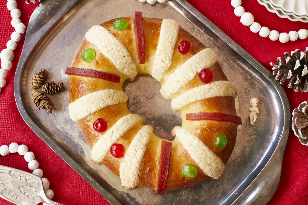 Rosca de Reyes (Three Kings Bread)is baked until golden brown. It's infused with orange zest and decorated with festive candied cherries, strips of quince paste, and sugar paste. A baby Jesus figurine is inserted into the bread after baking,