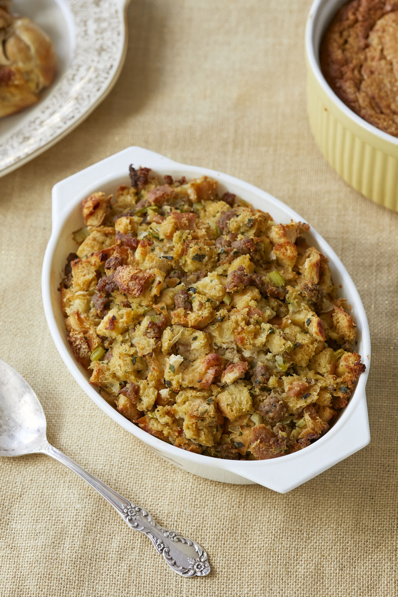 Classic Sage Sausage Stuffing is baked in a white milk galss baking dish. The dressing is alsos erved on a big golden-rim platter and a yellow baking dish. Toasted white bread is tossed with savory sausage, earthy sage, and aromatic onion and celery, mixed with in eggs and broth to a fluffy, moist casserole with tempting craggy browned bits!