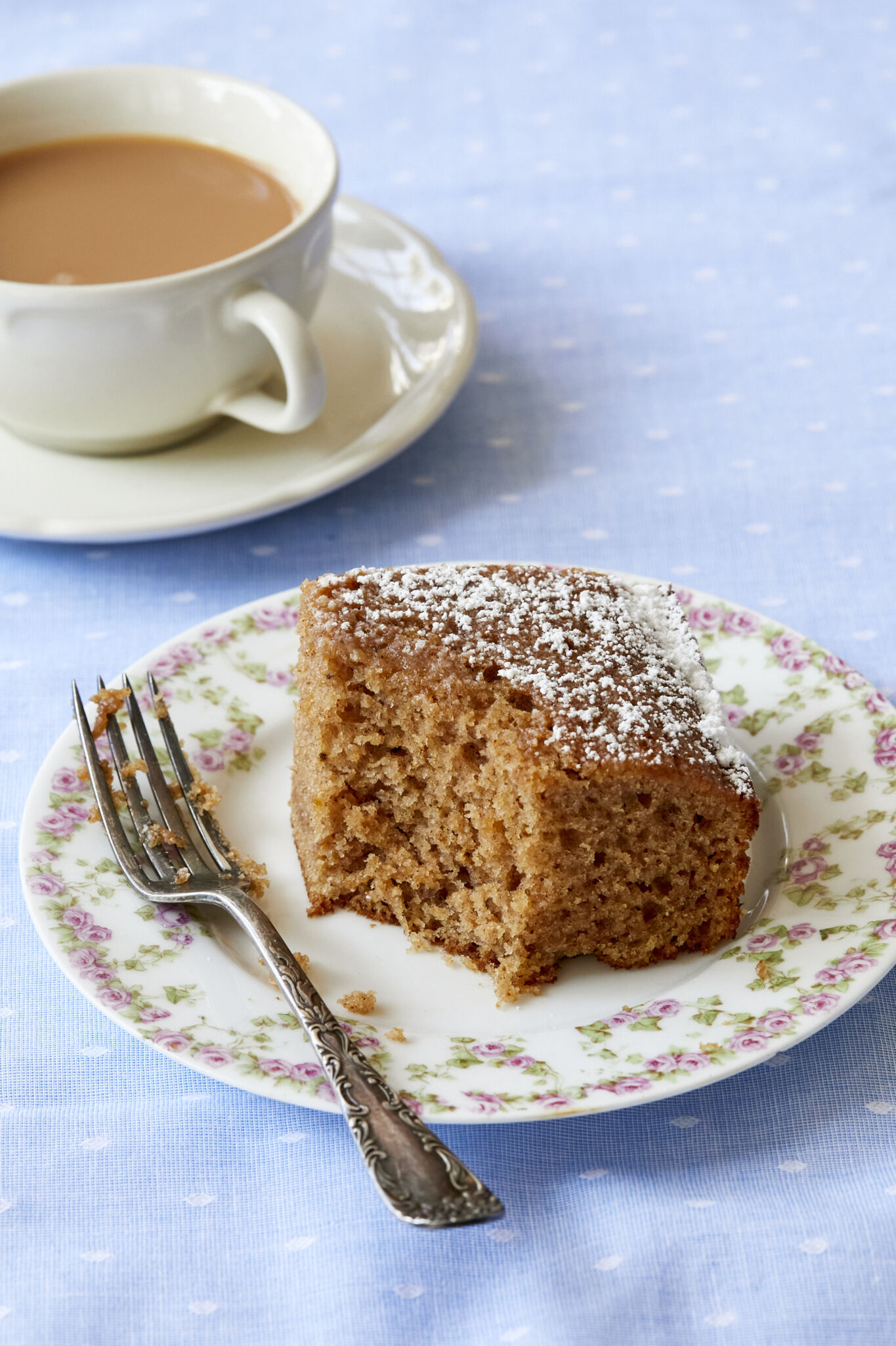 A generous square of Traditional Irish Spice Cake dusted with powdered sugar is served on a floral dessert plate. One bite has been taken and it clearly shows the delectably moist, fine crumb. The cake is paired with a cup of tea. 