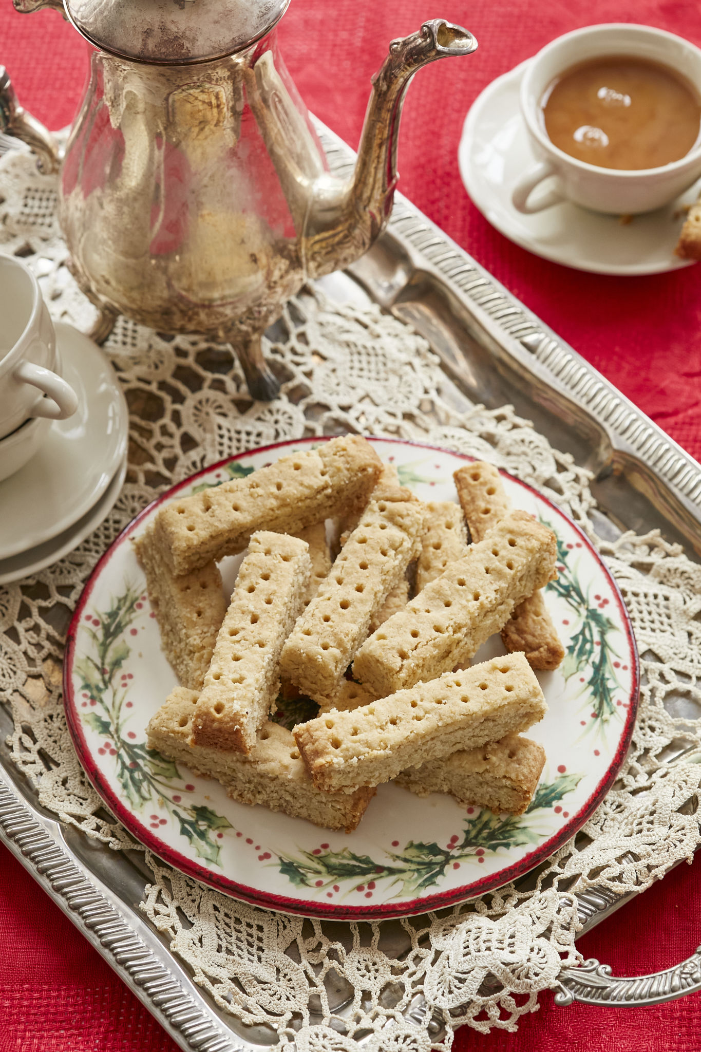 Crumbly Scottish shortbread cookies are served on a Christmas plate, decorated with holly berries, on a platter alongside two mugs and a silver decorative teapot.